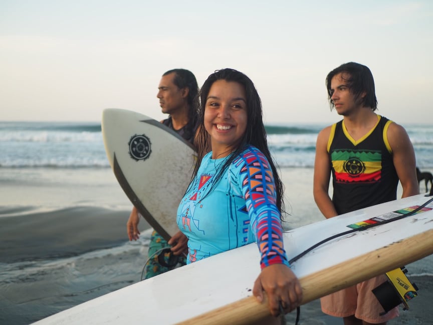 A group of young people carry surf boards on a beach and smile